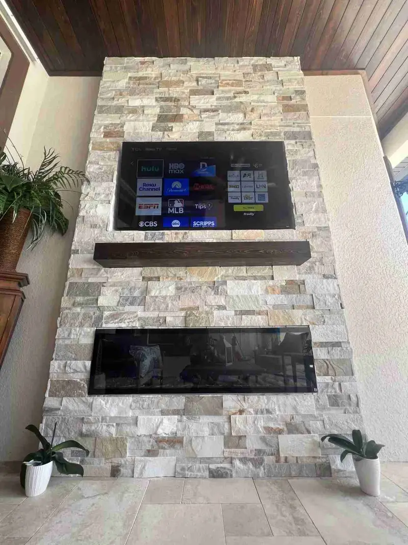 Ledgestone outdoor fireplace wall with a TV and a wood mantel on a covered lanai with a wood ceiling