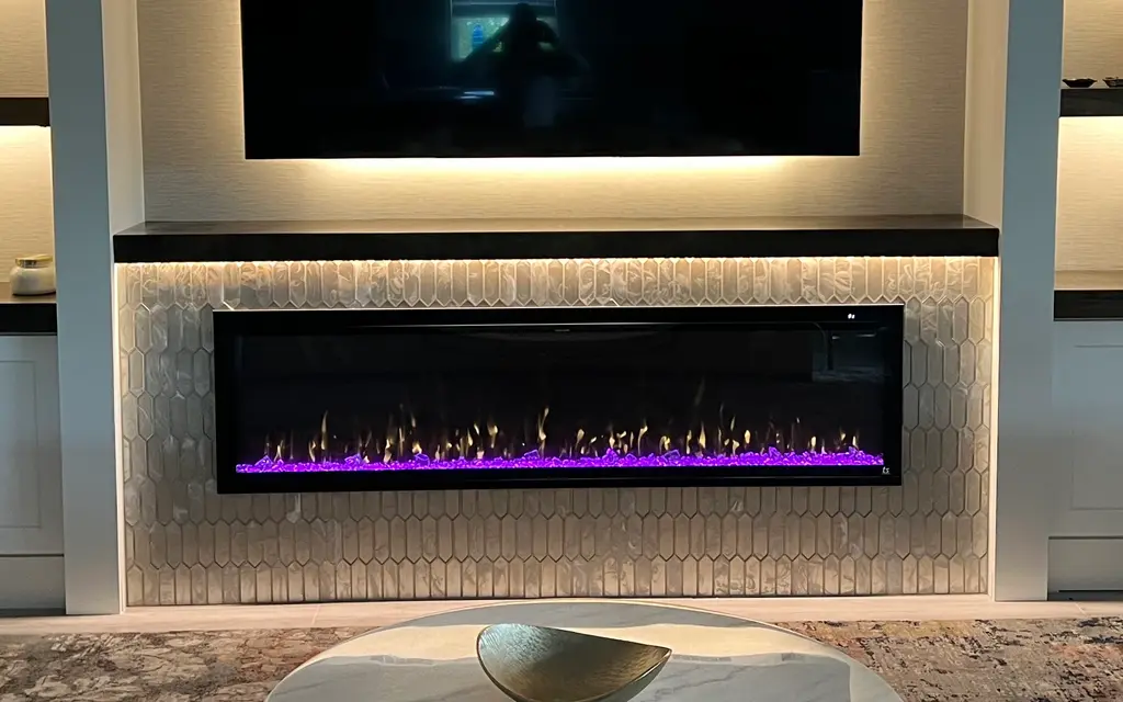 Linear electric fireplace set in a white tile surround below a TV, between lit built-in shelves